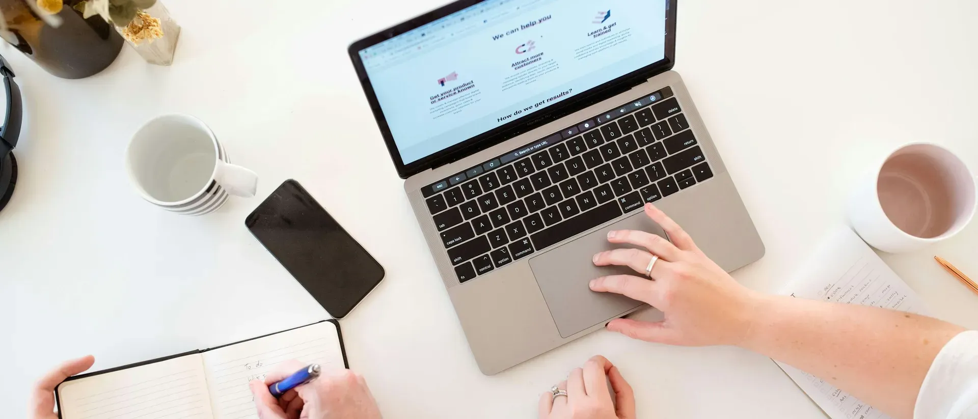 Twee collega's aan een wit bureau met een MacBook, een smartphone en een notitieboek, van bovenaf gezien
