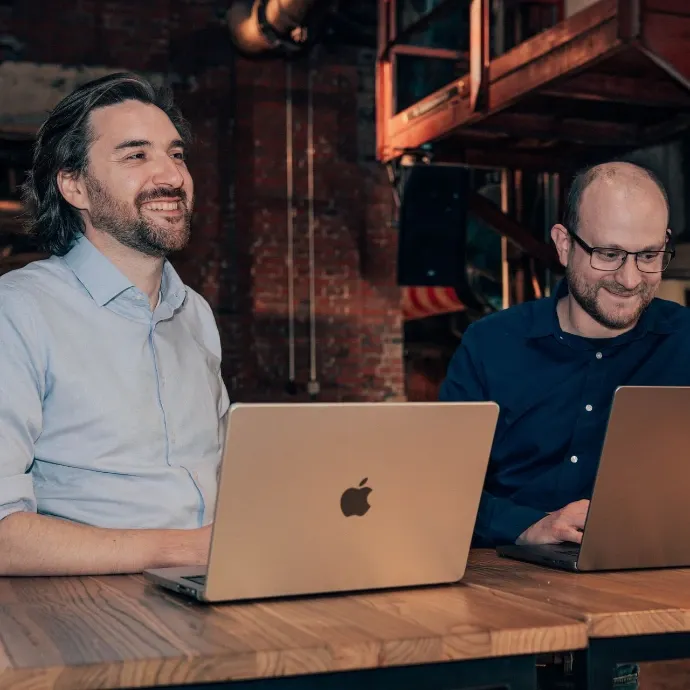 Raf en Gunther lachend aan een houten tafel, elk achter een MacBook