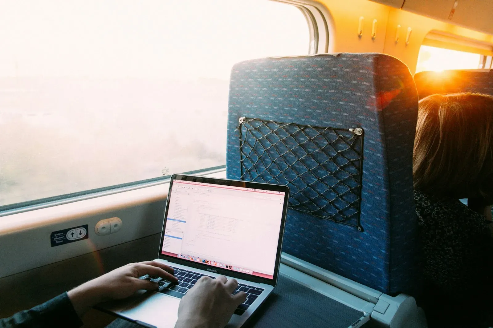 Iemand werkt op een MacBook aan het tafeltje van een trein, met laagstaande zon door het raam